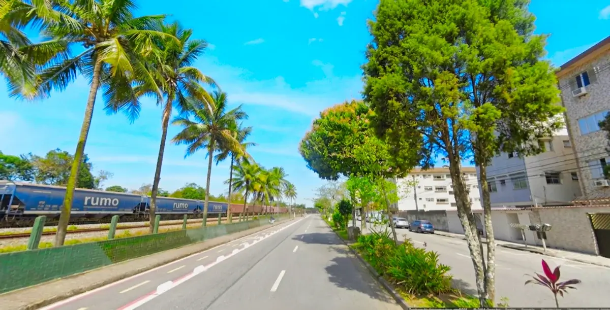 Avenida arborizada em Cubatão, com ciclovia, prédios e linha ferroviária com trem de carga passando, ao lado.