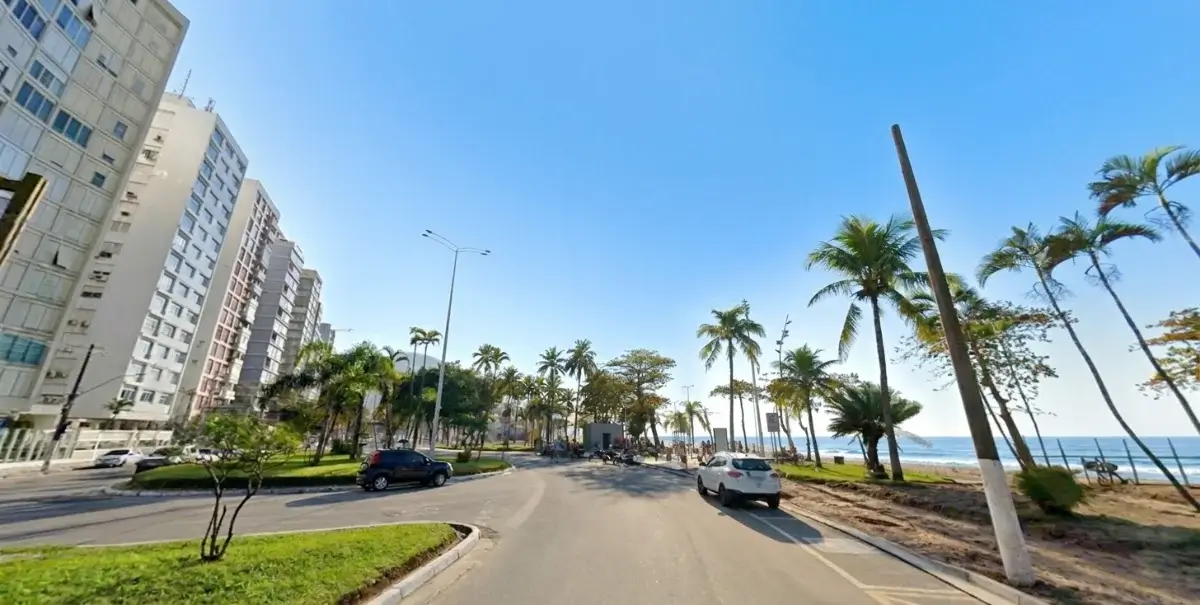 Avenida à beira-mar em São Vicente, com prédios, jardins, palmeiras e a praia ao fundo.